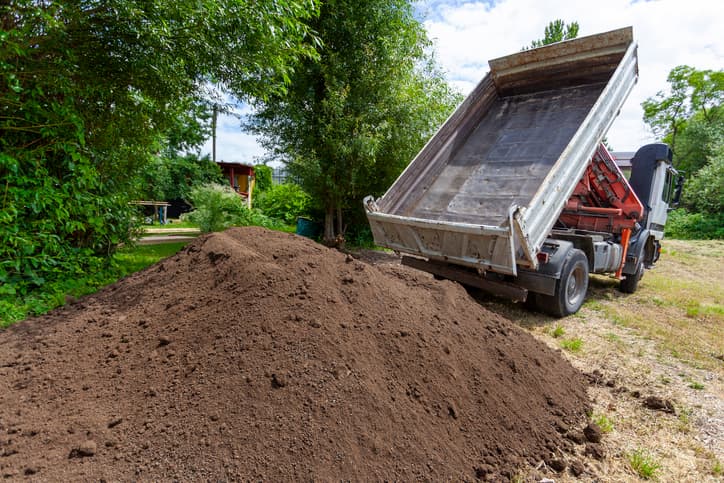 A dump truck is unloading a large quantity of brown earth into the backyard